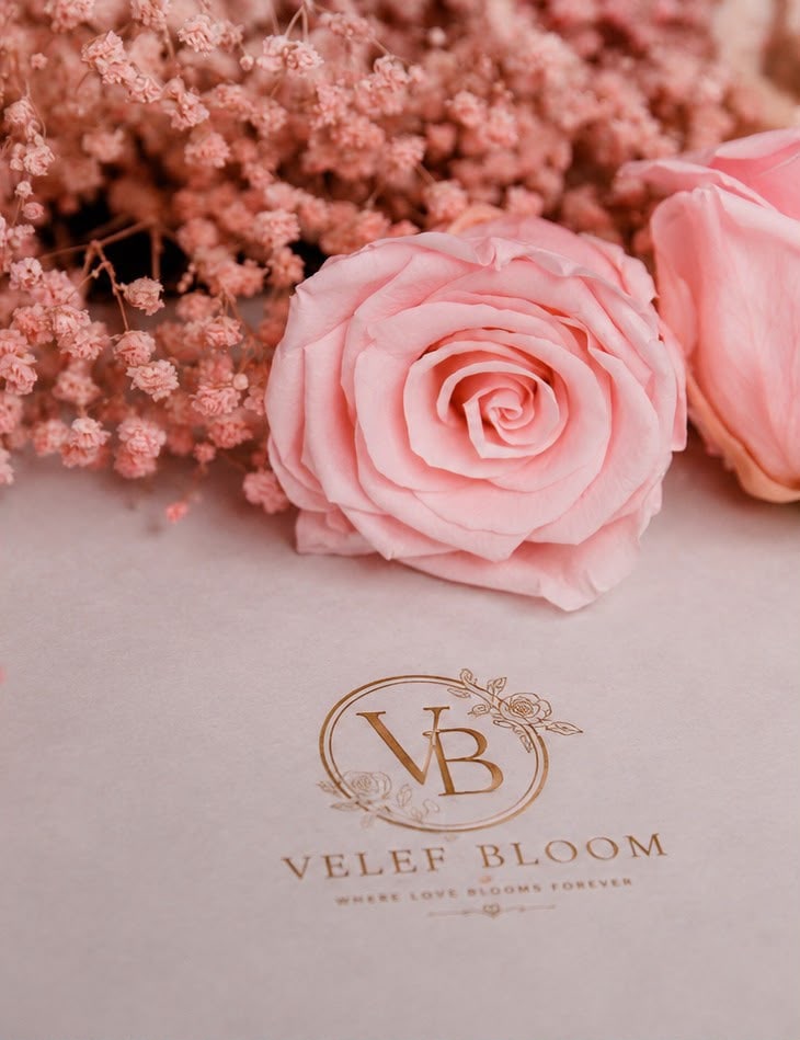 El monograma dorado de Velef Bloom y la frase Where Love Blooms Forever grabados sobre una superficie rosa, junto a una rosa preservada y nube seca